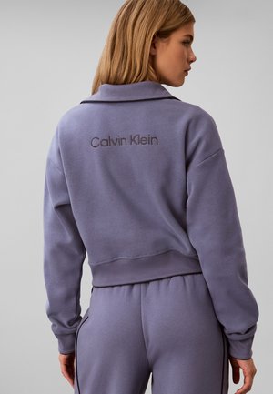 Γυναίκα που φορά μωβ φούτερ Calvin Klein και αντίστοιχο παντελόνι, εμφανιζόμενη από πίσω με απλό γκρι φόντο.
