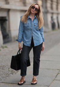 Camisa de ganga azul-claro com dois bolsos no peito, calças de ganga pretas de cintura alta, chinelos castanhos e uma mala de ombro preta. Óculos de sol estão usados.