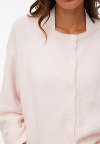 Gaiši rozā kardigans no mīksta, pūkaina materiāla ar apaļu izgriezumu kaklam, priekšpusē četri balti pogas un nolaisti pleci.