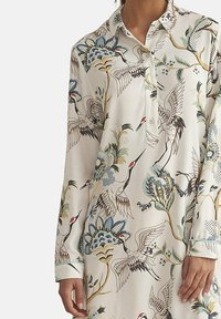 Chemise à manches longues en tissu crème avec un imprimé d'oiseaux et de fleurs en bleu, vert et marron, dotée d'un col et de poignets boutonnés.