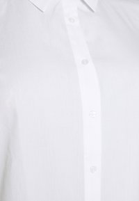 Vit skjorta med knappar framtill, gjord av lätt, strukturerad tyg med spetsig krage och små, genomskinliga plastknappar.