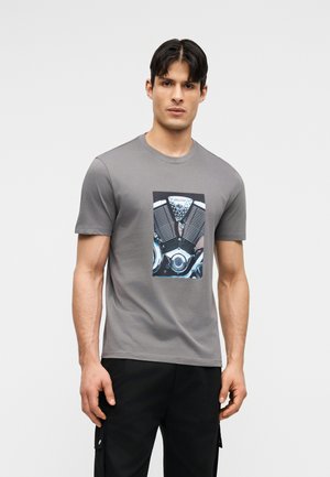 Άνδρας που φοράει γκρι μπλουζάκι με λεπτομερή γραφικό μοτοσυκλετιστής κινητήρα στο μπροστινό μέρος, συνδυασμένο με μαύρο παντελόνι, στέκεται μπροστά σε απλό φόντο.