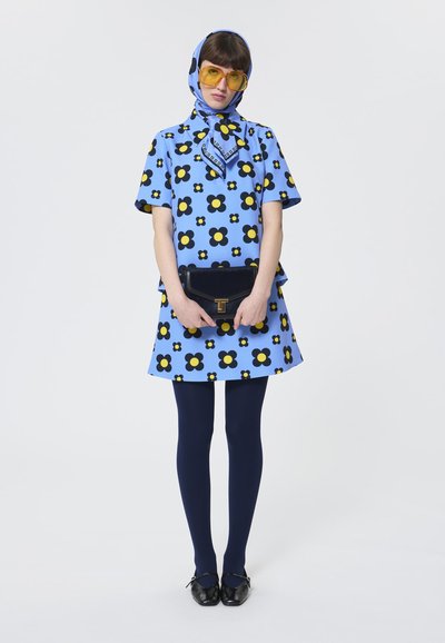 Blaues Blumenkleid mit schwarzen und gelben Blumen, kombiniert mit marinblauen Strumpfhosen und schwarzen Schuhen. Enthält einen passenden blauen Schal und übergroße gelbe Brille.