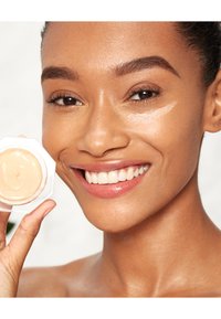 Clear cream in an octagonal jar held by a person. Smooth, reflective texture; warm beige color. Skin with a light, even coverage applied.