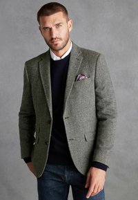Blazer gris à motif en chevrons avec deux boutons, porté sur un pull bleu marine et une chemise blanche à col, agrémenté d'un pochettede couleur.