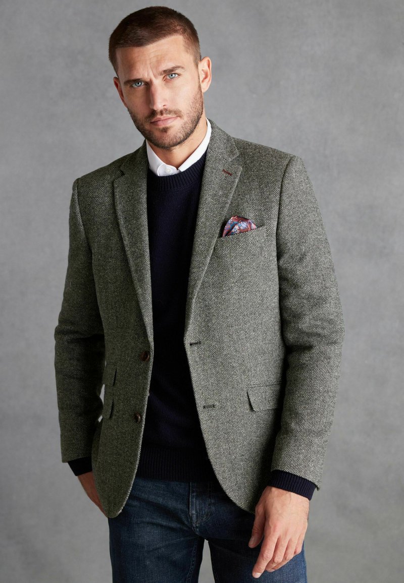 Blazer gris à motif en chevrons avec deux boutons, porté sur un pull bleu marine et une chemise blanche à col, agrémenté d'un pochettede couleur.