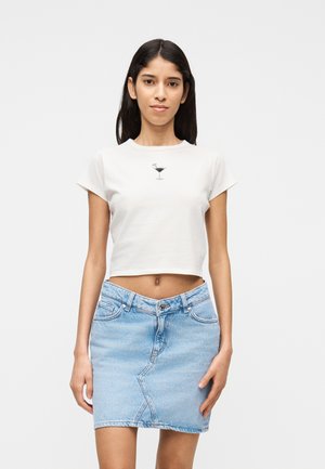 Ung kvinde med langt mørkt hår iført en hvid kort top med print af et cocktailglas og en lyseblå denimnederdel.