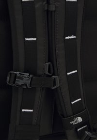 Gros plan sur des bretelles noires de sac à dos avec boucle en plastique réglable et couture blanche ; logo The North Face visible sur une bretelle.