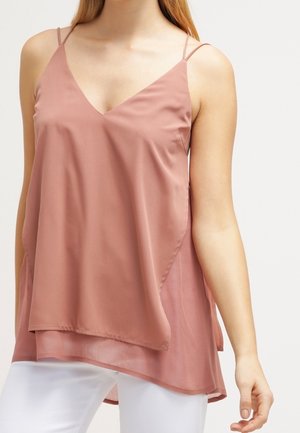 Frau trägt ärmelloses, staubrosafarbenes, geschichtetes Tunika-Top mit Spaghettiträgern und weiße Hose vor einfachem Hintergrund.