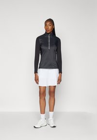 Černý dlouhý rukáv s límcem na půl zip, v kombinaci s bílou sukní. Model má na sobě bílé sportovní boty a pono�žky. Jednoduchý design.