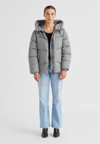 Graue Puffy-Jacke mit Kapuze, die eine strukturierte Außenseite, ein gepolstertes Design, einen Reißverschluss und zwei Fronttaschen aufweist. Wird über einem schwarzen Oberteil getragen.