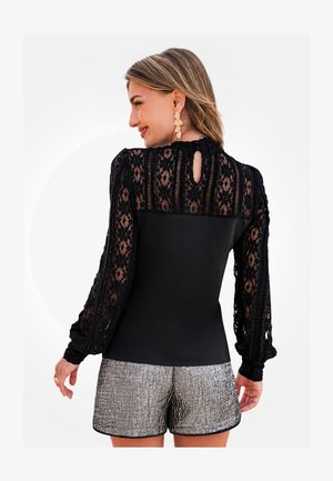 Schwarzes Oberteil mit langen Spitzenärmeln und einem Ausschnitt im Rücken, kombiniert mit texturierten, metallisch silbernen Shorts.
