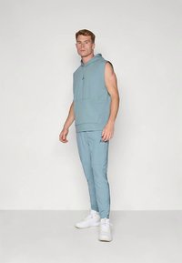 Vyras stovintis mėtos spalvos be rankovių džemperyje su kapišonu ir derančiose sportinėse kelnėse, dėvintis baltus sportbačius, priešais baltą tuščią foną.