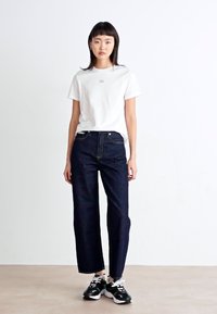 Jeans in denim blu scuro con taglio dritto, abbinati a una t-shirt bianca in cotone semplice. Il modello indossa sneakers nere e bianche.