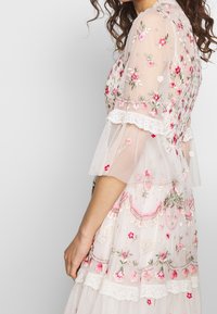 Femme portant une robe blanche transparente brodée de fleurs avec des détails roses et verts et une bordure en dentelle aux manches courtes en cloche.