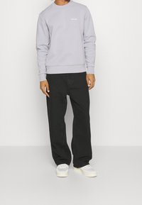 Sudadera de algodón gris claro con cuello redondo y logo de Calvin Klein en la parte superior izquierda. Pantalones holgados negros y zapatillas blancas.
