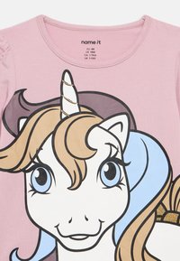 Ροζ μπλουζάκι για νήπια με ένα μεγάλο χαμογελαστό πρόσωπο μονόκερου που διαθέτει μπλε μάτια, καστανό και μωβ χαίτη, και ένα λευκό κέρατο με χρυσές γυαλιστερές λεπτομέρειες.