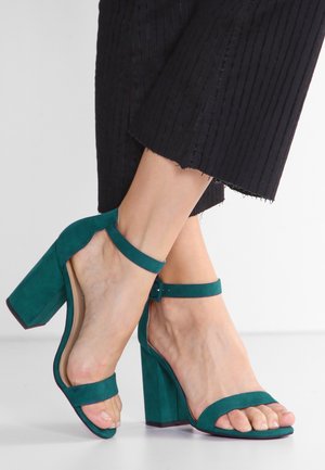 Teal suede high-heeled sandals with a wide strap over the toes, adjustable ankle strap, and block heel featuring a textured sole.