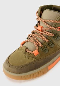 Botte de randonnée vert olive avec accents orange, dessus en suède texturé et mesh, semelle en caoutchouc, et lacets à motifs avec œillets pour un ajustement sécurisé.