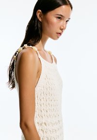 Top in cotone all'uncinetto di colore bianco, caratterizzato da un motivo di punti aperti. I dettagli notevoli includono perline beige sulle spalline, che accentuano il design.