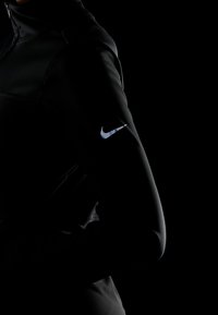 Sort sportjakke med høj krave, der har en glat, glansfuld tekstur og et reflekterende sølvfarvet Nike-logo på venstre ærme.