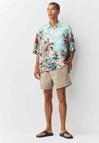 Kortærmet, turkiset tropisk skjorte med print af røde og grønne palmer, parret med beige shorts og sorte klipklappere.