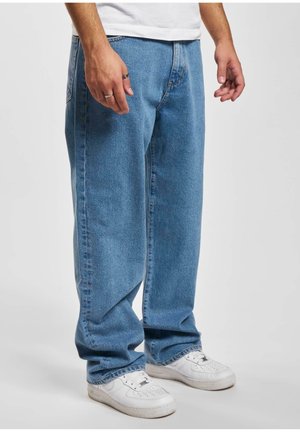 Mężczyzna w luźnych niebieskich dżinsach i białych trampkach stoi z rękami swobodnie opuszczonymi po bokach na szarym tle.