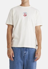 Camiseta de algodón color crema con mangas cortas, con un diseño impreso en rojo y azul en el pecho, corte relajado y cuello redondo.