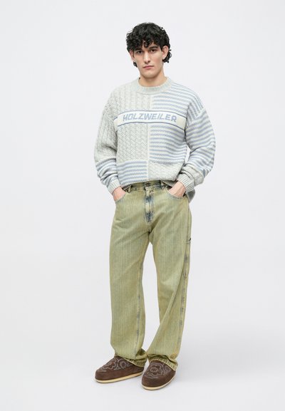 Szürke és világoskék kötött pulóver köteletmintával és HOLZWEILER felirattal, világoszöld csíkos farmerral és barna bebújós cipővel párosítva.