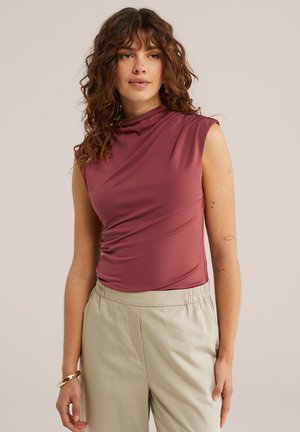 Femme aux cheveux bruns bouclés, portant un haut mauve sans manches et un pantalon beige, debout devant un fond clair uni.