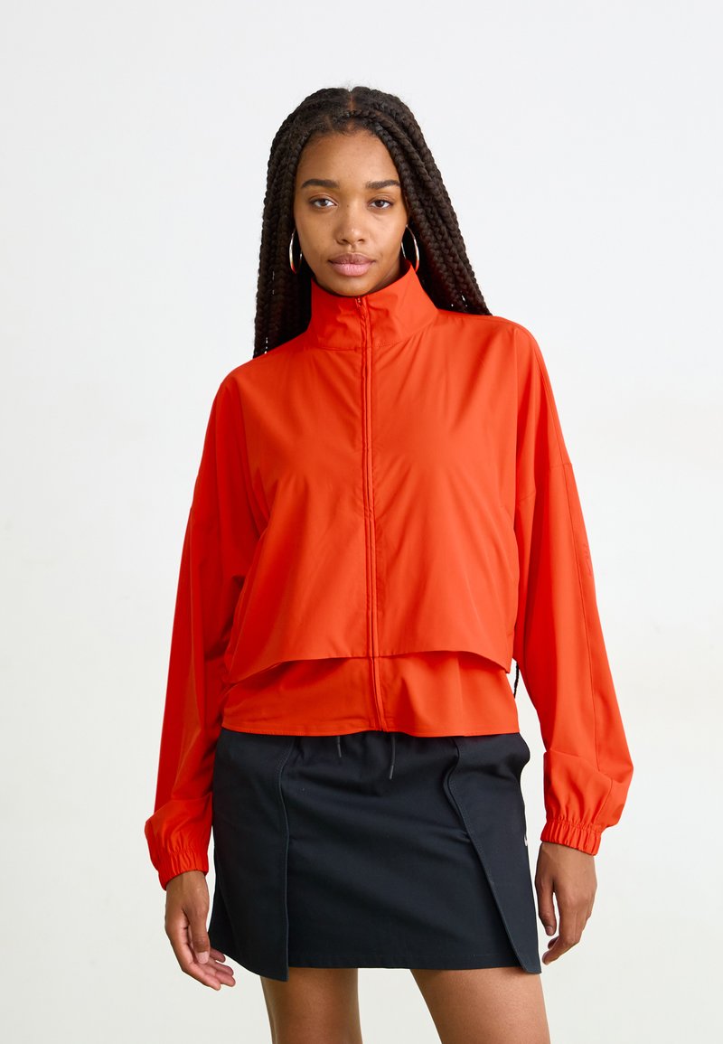 Rød zip-up jakke med høy krage, løs passform og elastiske mansjetter, parret med et svart skjørt med sidesliss og snøring.