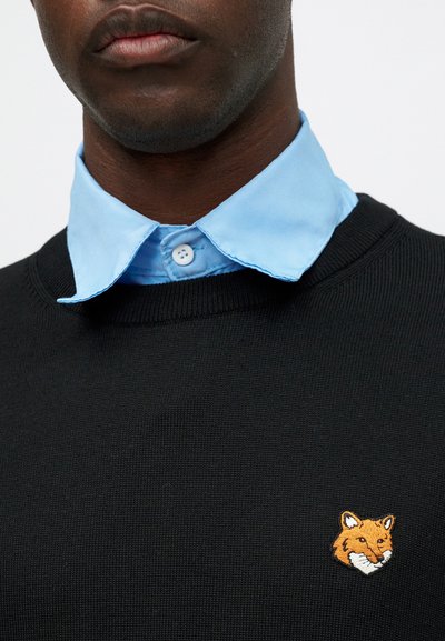 Nærbilde av en person som har på seg en svart genser med en brodert oransje revhode og en lyseblå skjorte under.