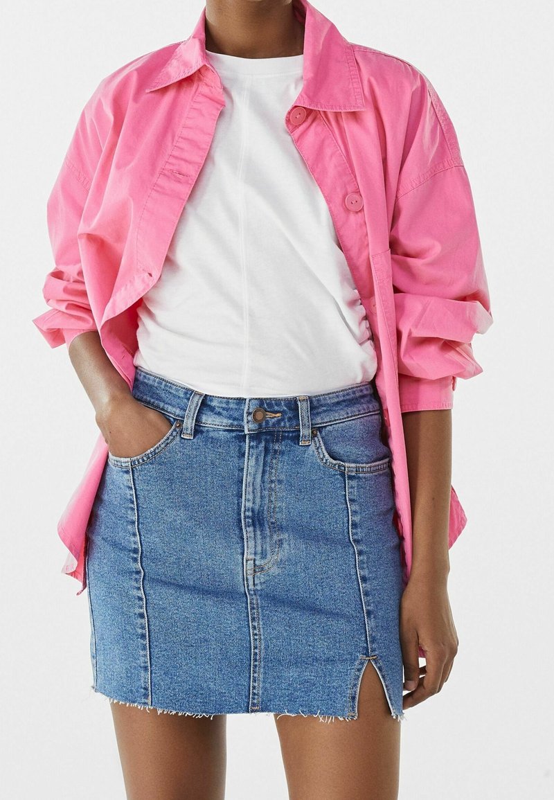 Světle růžová košile na knoflíky přes bílém tričku, spárovaná s modrou denimovou sukní s předním rozparkem a ošoupaným lemem.
