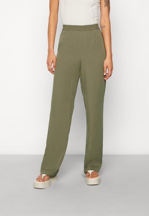 Femme portant un pantalon large à taille élastique olive, des sandales à plateforme blanches et un haut blanc sans manches, debout contre un mur blanc.
