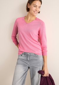 Roze langwerpige V-hals trui van lichtgewicht stof, gecombineerd met lichtgrijze jeans. De persoon houdt een kastanjebruine jas in één hand.