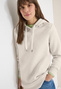 Jeune femme portant un sweat à capuche beige clair et une chemise verte, la main dans la poche, appuyée contre un mur blanc, souriant doucement.