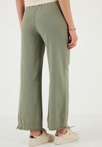 Hellgrüne weit geschnittene Hose aus strukturiertem Stoff, mit fransigen Säumen und einem elastischen Bund, kombiniert mit beigen Sneakers mit roségoldenen Akzenten.
