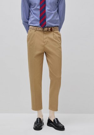 Uomo che indossa pantaloni beige plissettati, una camicia blu a quadri, una cravatta a righe rossa e blu, una cintura beige, calzini chiari e mocassini neri, in piedi su sfondo semplice.