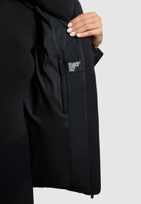 Sorte pufferjakke med en skinnende overflade, der har et sømt logo-patch på det indre for og et elegant, minimalistisk design.