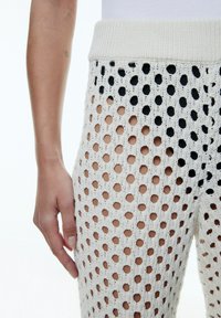 Gebreide broek in crème met een groot open-holes patroon, voorzien van een geribde tailleband. Het ontwerp is lichtgewicht en ademend, waarbij de huid eronder zichtbaar is.