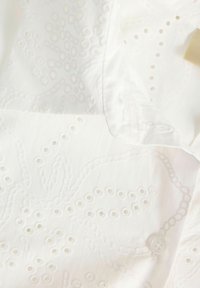 Λευκό ύφασμα με περίτεχνη κεντητική με τρυπητό μοτίβο και ραμμένο λευκό κουμπί σε ένδυμα.