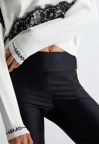 Weißer Pullover mit schwarzem Spitzenbesatz und Fransen, kombiniert mit hochtaillierten schwarzen Leggings. Der Pullover hat ein Logo am Ärmel.