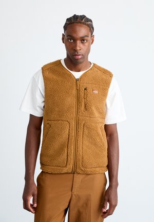 Young man with braided hair wearing a tan fleece zip-up vest over a white t-shirt and matching tan pants, standing against a plain background.