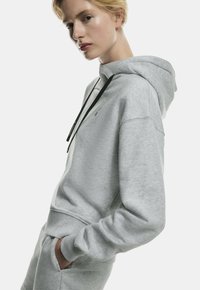 Gray hoodie made of soft material, featuring a kangaroo pocket, black drawstring, and a subtle logo on the chest. Textured fabric finish.