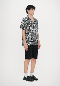 Μπλουζάκι κοντομάνικο με μαύρο και άσπρο μοτίβο και αφηρημένα σχήματα, σε συνδυασμό με μαύρες βερμούδες και μαύρες λαστιχένιες παντόφλες, φορεμένο με άσπρες κάλτσες.