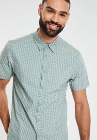 Kurzarmpullover mit Knopfleiste in hellgrün mit einem kleinen weißen Blumenmuster, aus einem glatten Stoff gefertigt. Hat einen klassischen Kragen und Knöpfe.