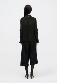 Blusa preta transparente com mangas longas e detalhes em babados, combinada com calças pretas de perna larga. Usada com sapatos de salto alto pretos.