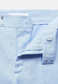 Pantalon en lin bleu clair avec une fermeture à boutons, une ouverture de poche horizontale, et des accents métalliques sur la ceinture. Texture lisse et design épuré.