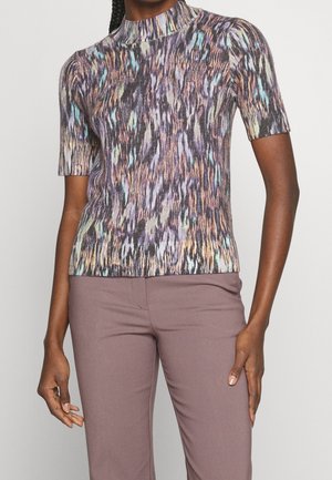 Kobieta ubrana w krótki top z wysokim kołnierzem o wielokolorowym abstrakcyjnym wzorze oraz dopasowane spodnie w kolorze wrzosowym, stojąca na jednolitym tle.