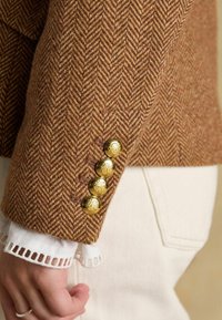 Blazer călduț de culoare maro cu un model herringbone, decorat cu butoane aurii și un manșon din dantelă albă care iese de sub acesta. Textura materialului este aspră și texturată.